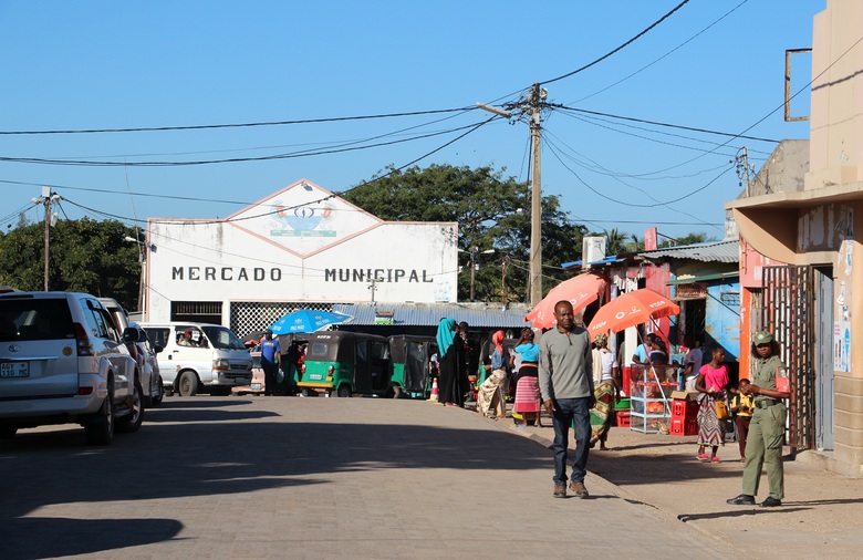 Straßenszene im mosambikanischen Vilankulo. Eine Straße, auf der Menschen zu Fuß unterwegs sind, führt auf eine Markthalle zu.